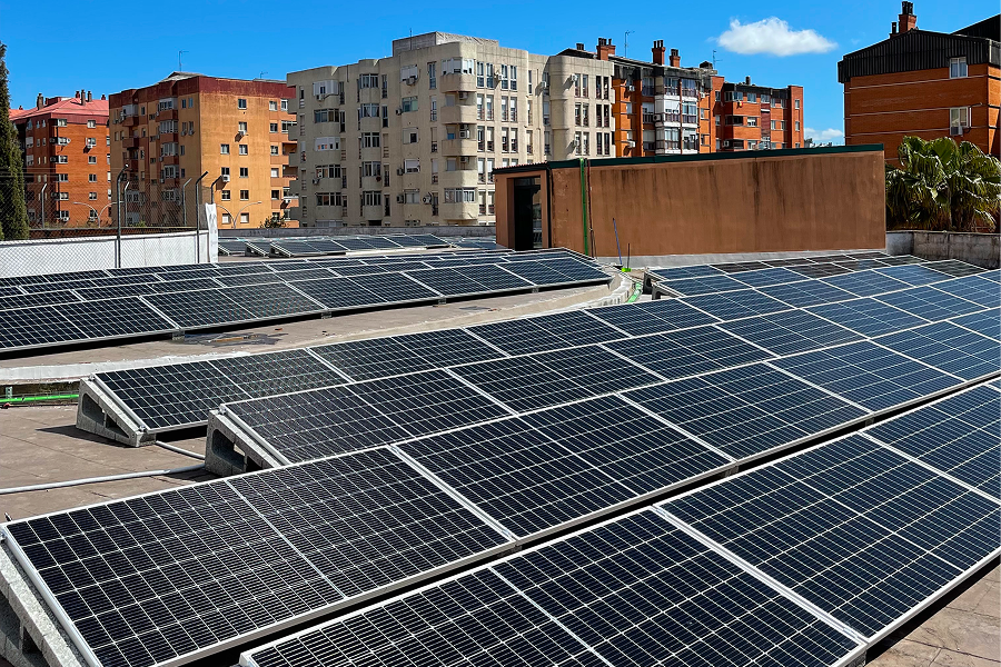 Vista de paneles solares instalados en un tejado, con una fila de edificios de apartamentos de fondo. El cielo es de un azul claro con algunas nubes dispersas.