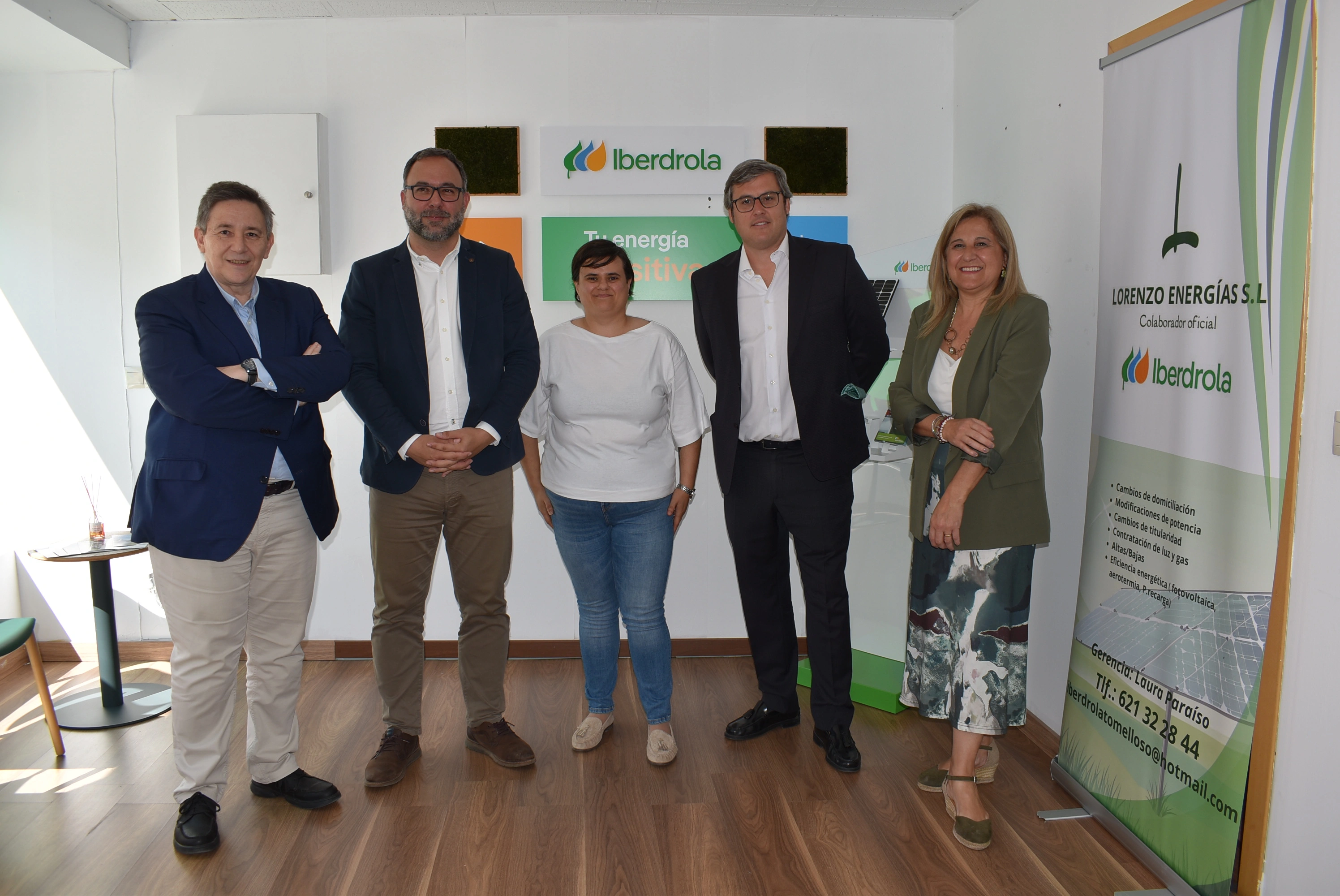 Grupo de personas de pie en un espacio de oficina, con un banner de Iberdrola al fondo. Se muestra un hombre con chaqueta azul, un hombre con blazer blanco, una mujer con camiseta blanca, un hombre con blazer negro y una mujer con blusa verde. Todos están de pie sonriendo y en un ambiente luminoso.