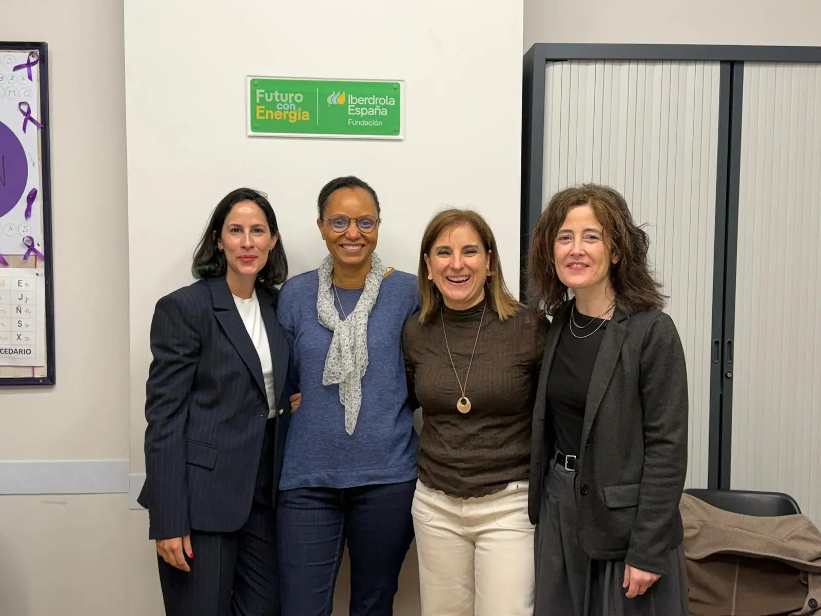 Four women posing together in a workroom. The background wall features a sign that reads "Futuro con Energía" along with the Iberdrola logo. The women are dressed professionally in suits and sweaters. They are all smiling while slightly embracing each other.