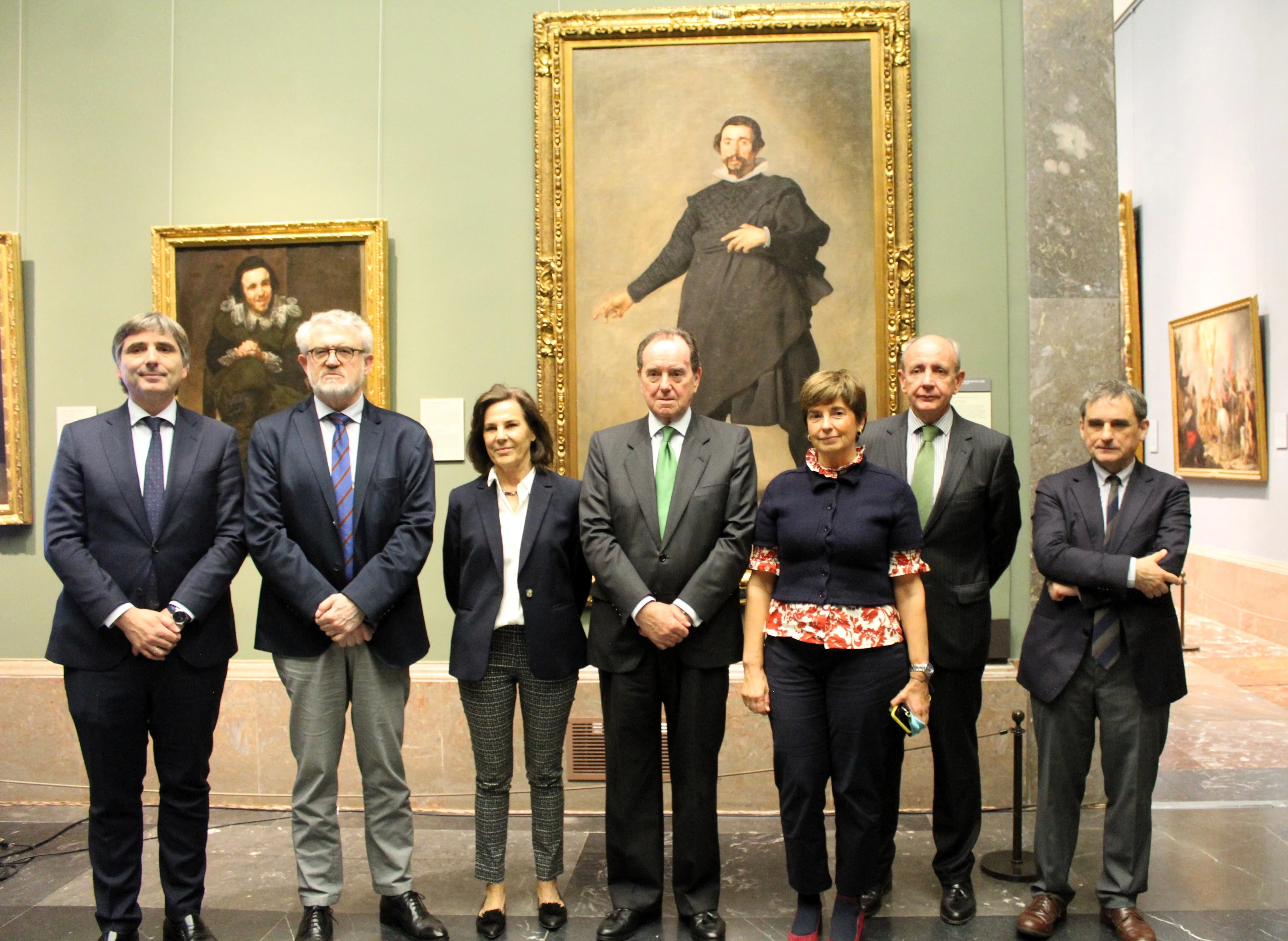 A group of seven people posing in front of a portrait in an art gallery. The artwork in the background shows a man in historical attire. The group members are dressed formally, with suits and shirts, and some wear green ties. The scene is well-lit and decorated with additional paintings on the walls.