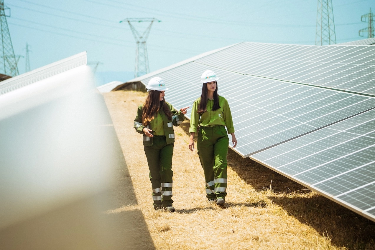 Two women in green uniforms and safety helmets walking next to solar panels in a field, talking while observing the landscape.