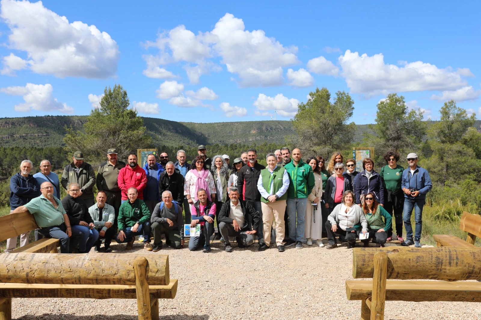 Group of people posing outdoors in front of a mountainous landscape with vegetation and a blue sky with clouds.