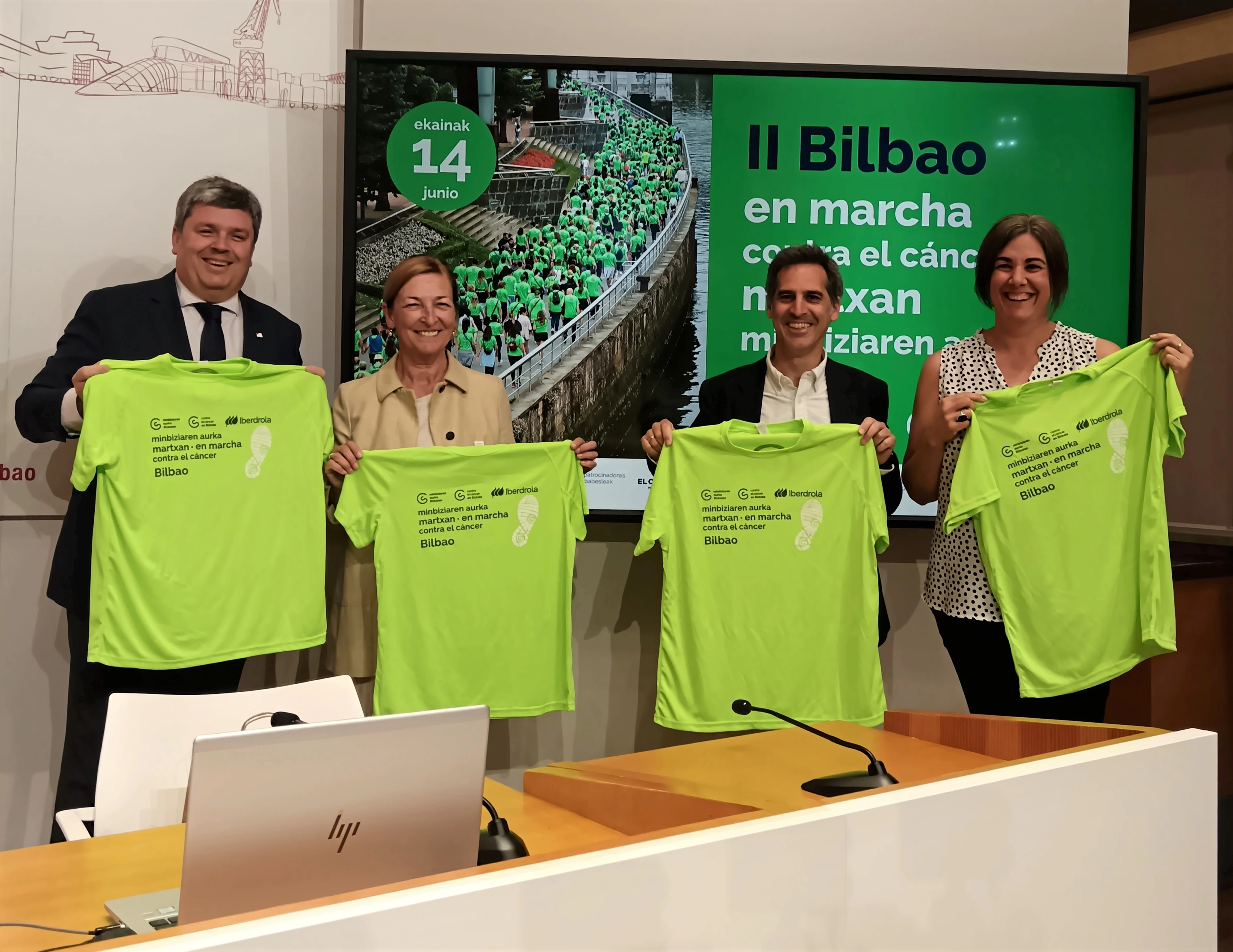 Cuatro personas posando en una rueda de prensa sosteniendo camisetas verdes del evento “II Bilbao en Marcha contra el cáncer”, con cartel informativo al fondo.