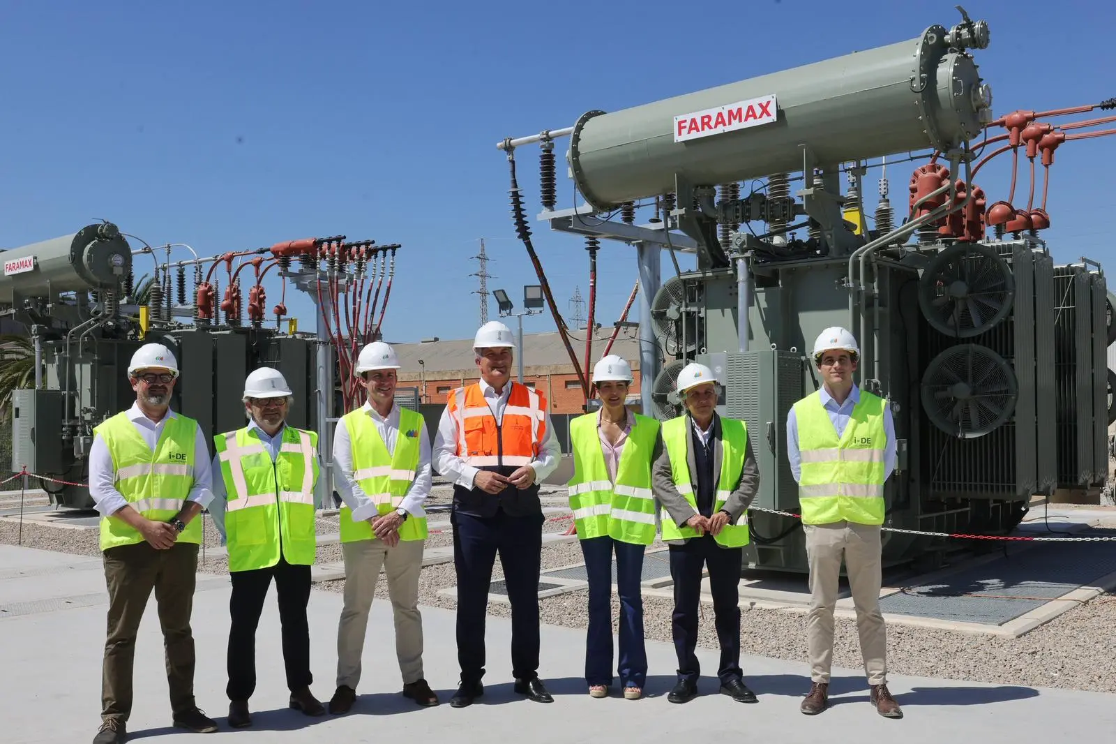 Siete personas posan frente a transformadores eléctricos de gran tamaño en una subestación al aire libre. Todas llevan casco de seguridad blanco y chaleco reflectante, seis de ellos verde y uno naranja. Los transformadores son grises con cables de alta tensión conectados, y uno tiene un cartel que dice "FARAMAX". El cielo está despejado y azul.