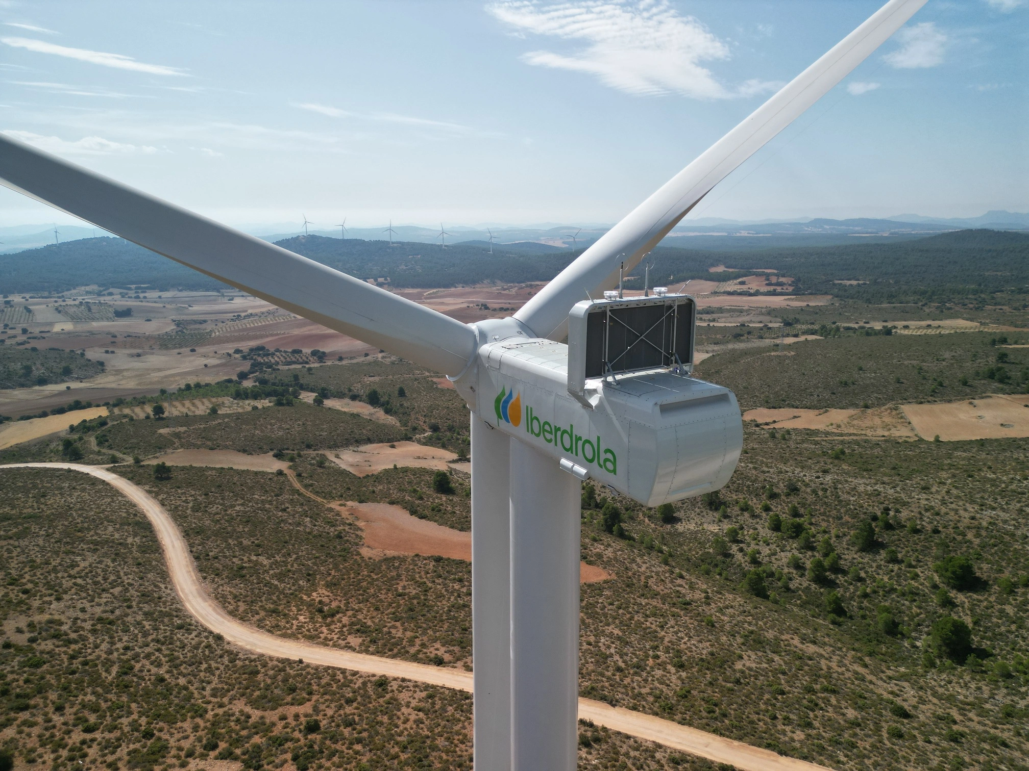 Primer plano de la góndola y las aspas de un aerogenerador de Iberdrola en un parque eólico, con un paisaje de colinas y otros aerogeneradores al fondo.