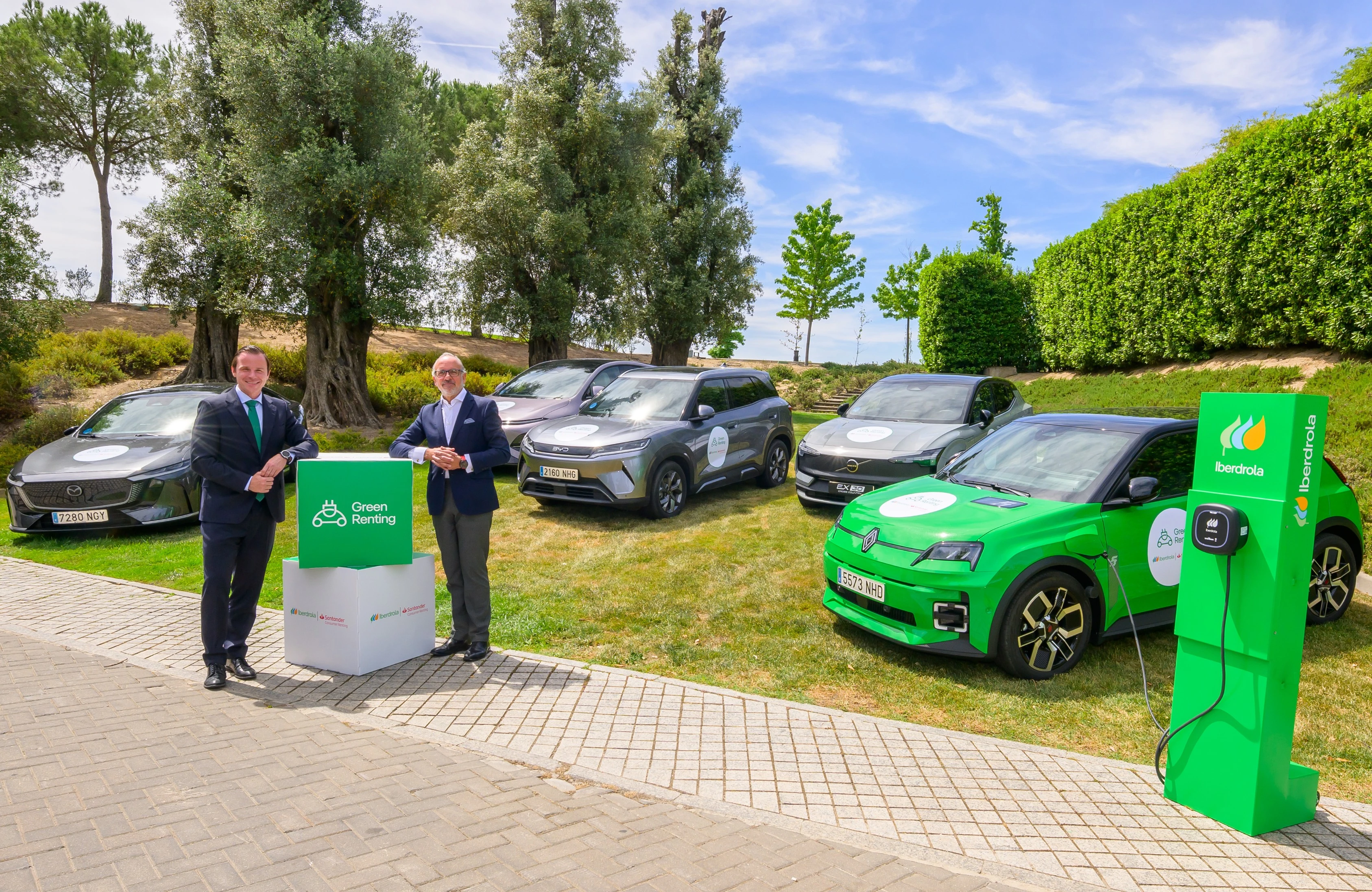 Dos hombres posando junto a vehículos eléctricos de Iberdrola en un espacio al aire libre, con estaciones de carga verde y arbustos de fondo, en la presentación del proyecto Green Renting.