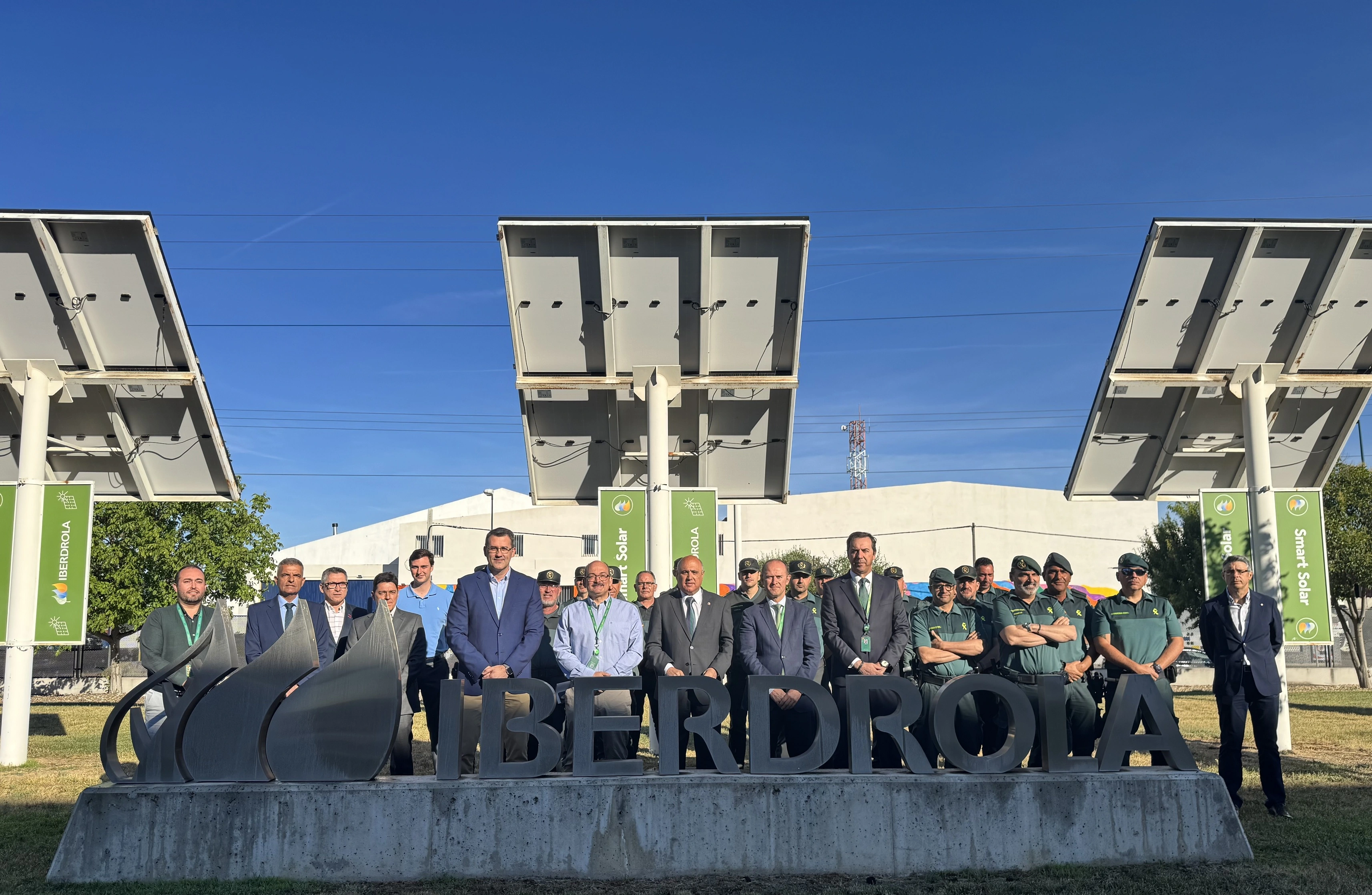 Group of Iberdrola employees and Civil Guard posing in front of solar panels and Iberdrola logo under clear sky.