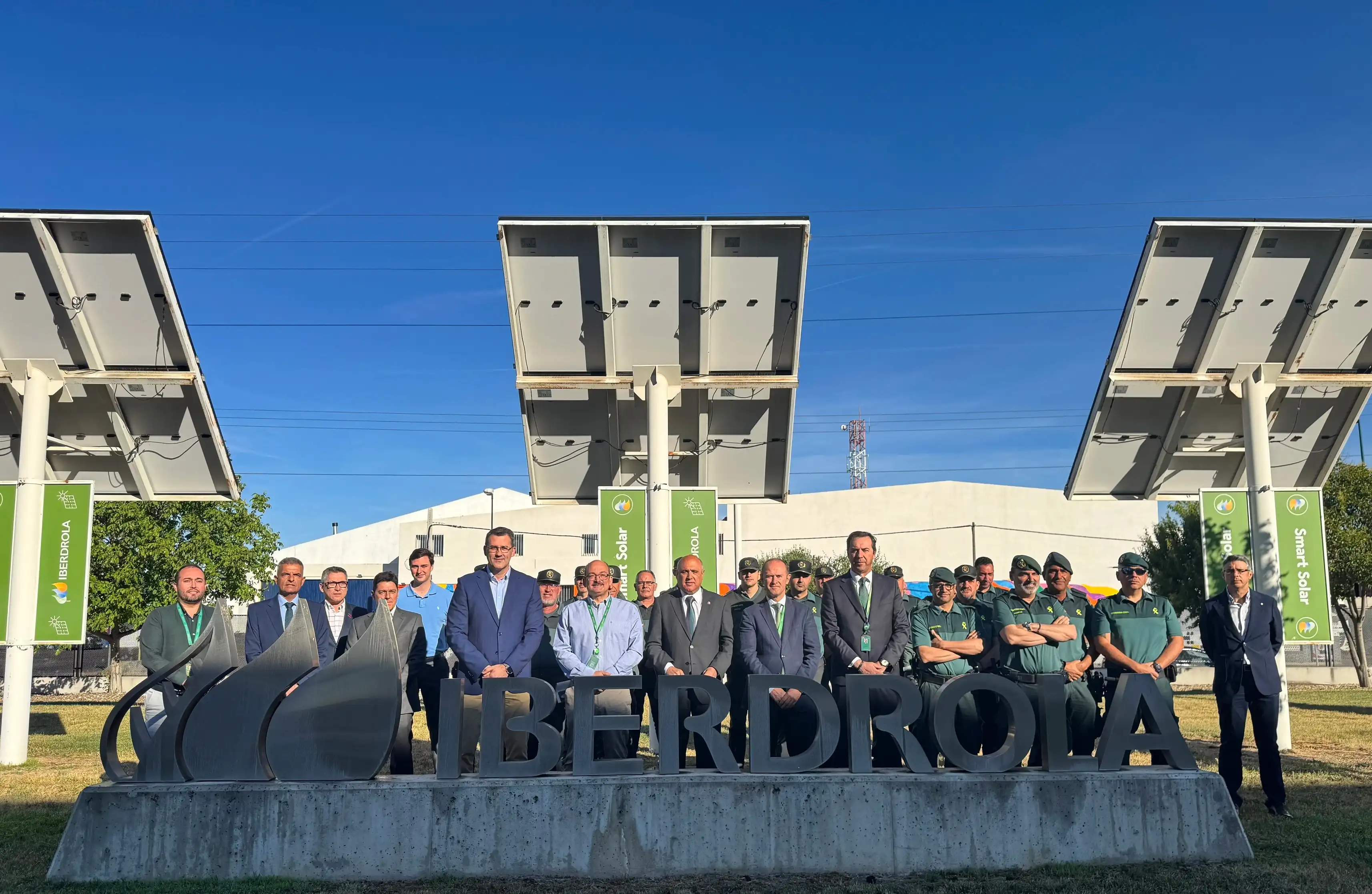 Group of Iberdrola employees and Civil Guard posing in front of solar panels and Iberdrola logo under clear sky.