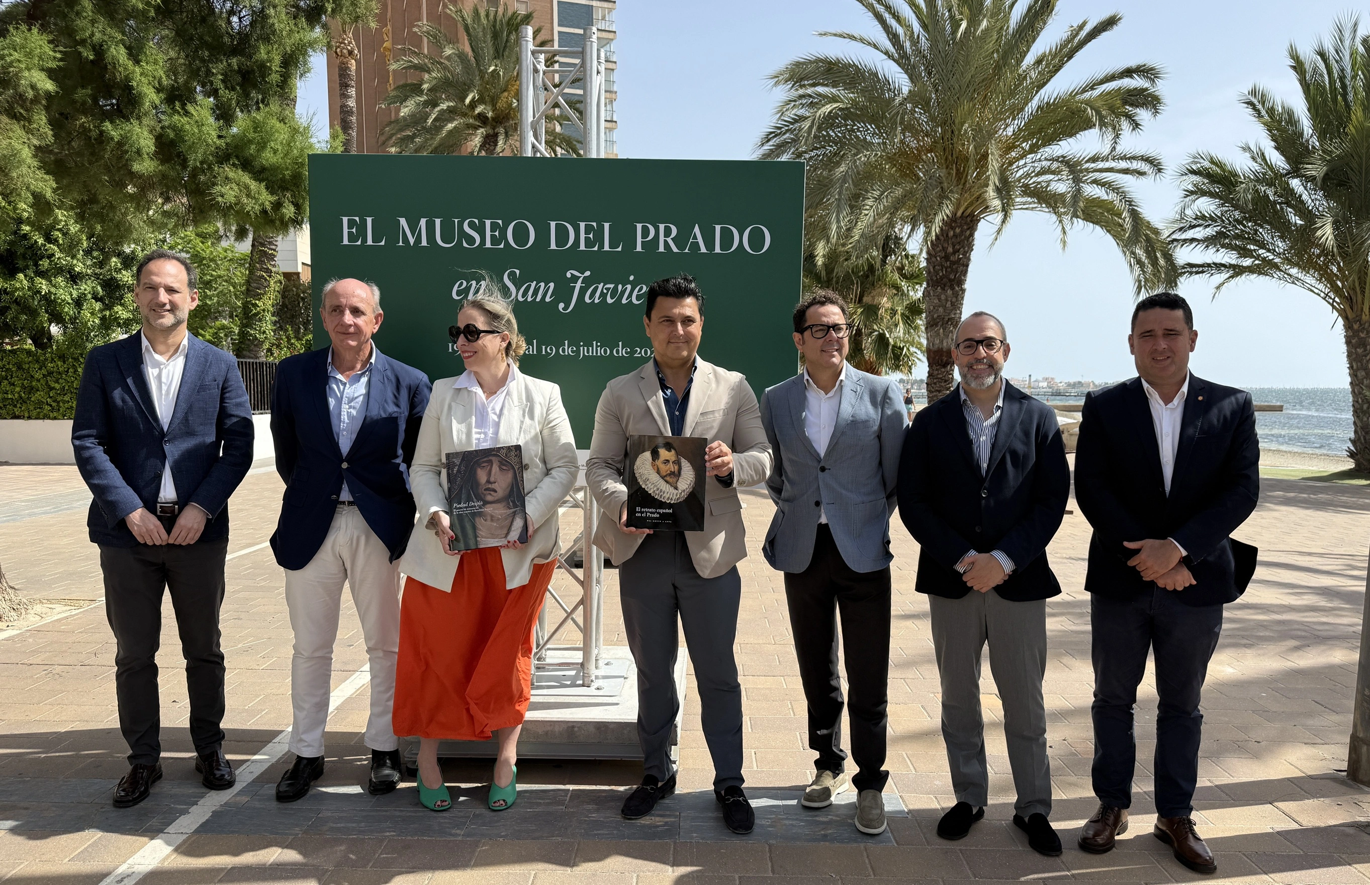 Grupo de representantes institucionales posando en el exterior junto a un panel del Museo del Prado en San Javier, en un acto cultural.