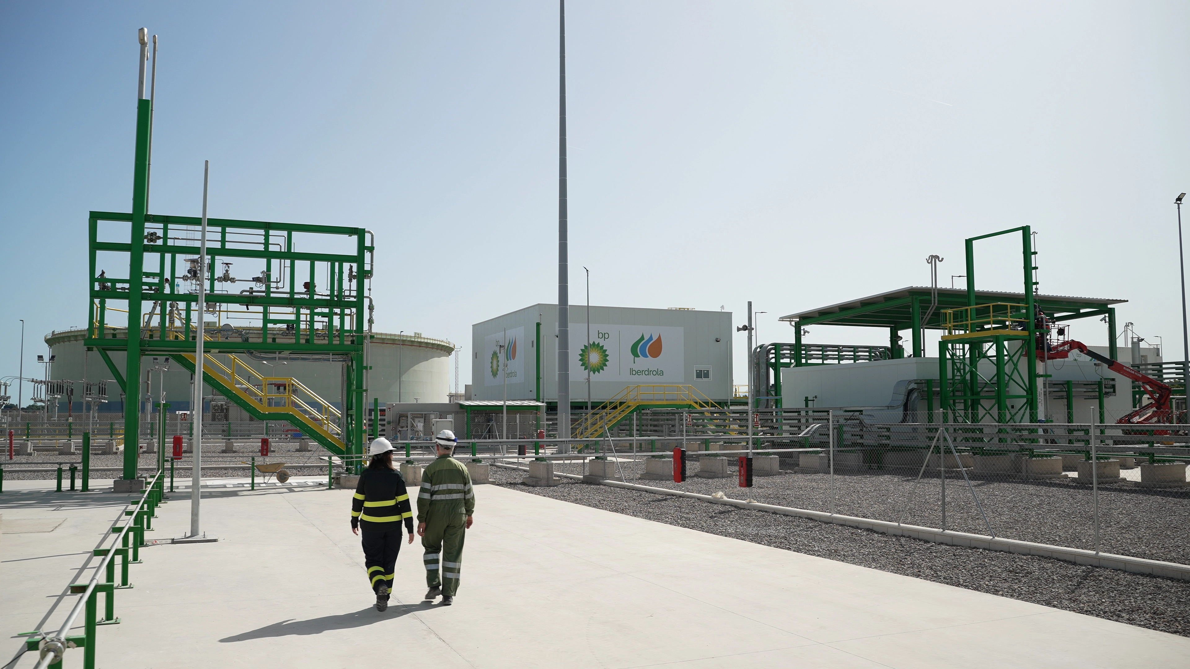 Two workers in protective equipment walking through green hydrogen plant with industrial structures, pipelines and storage tanks.
