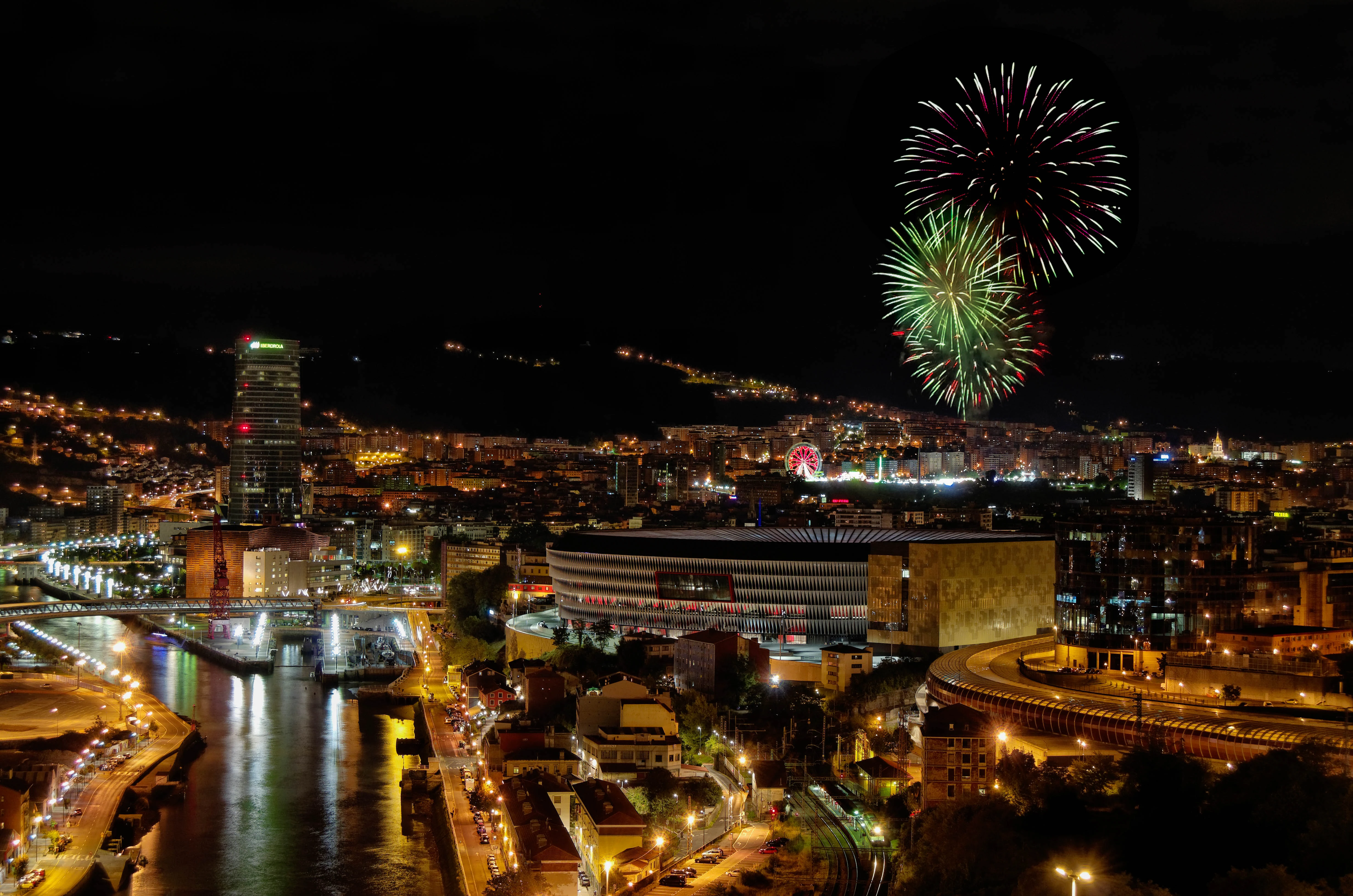 Vista nocturna de Bilbao con fuegos artificiales sobre la ciudad y la Torre Iberdrola iluminada.