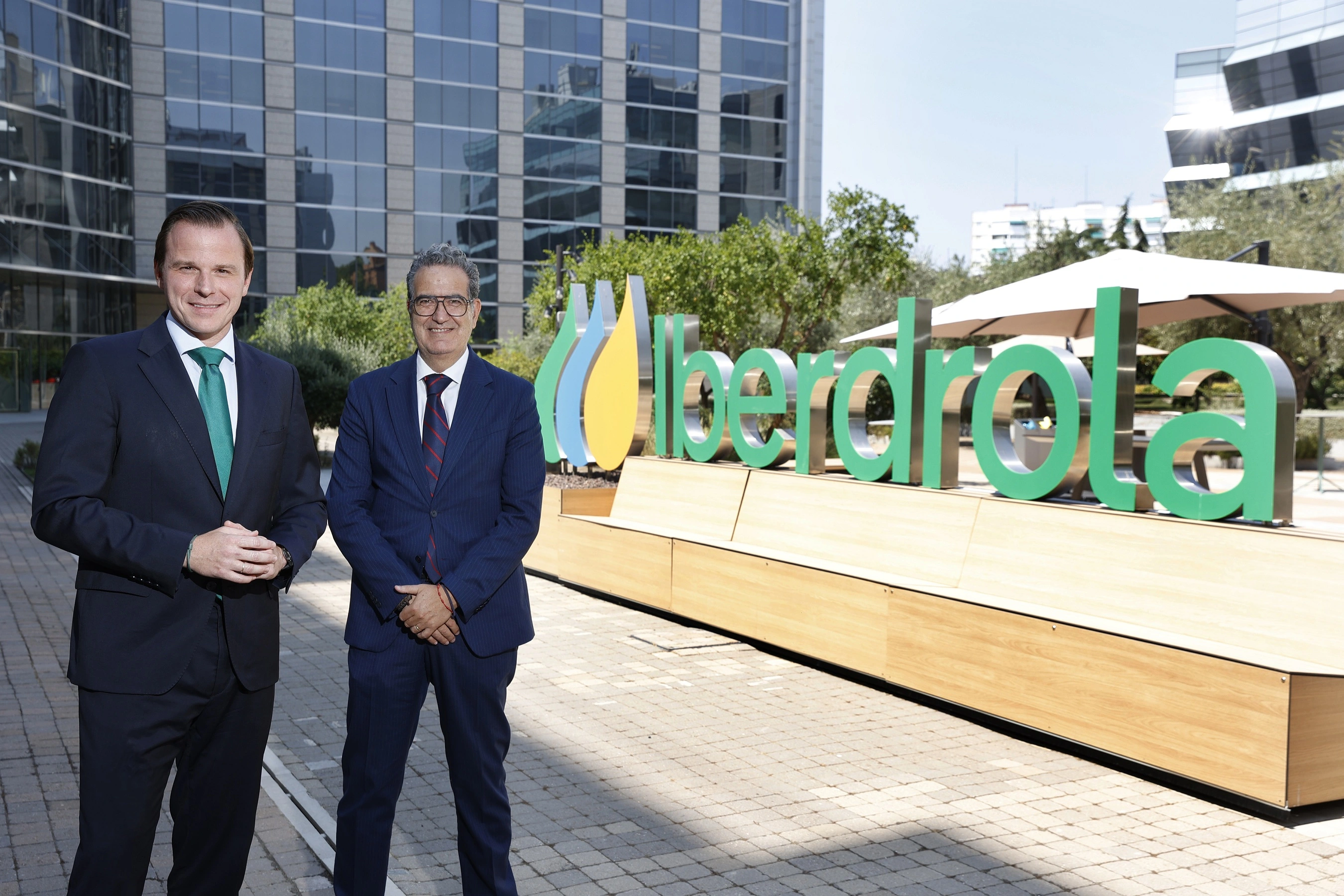 Dos directivos posan junto al logotipo de Iberdrola frente a un edificio de oficinas corporativas.