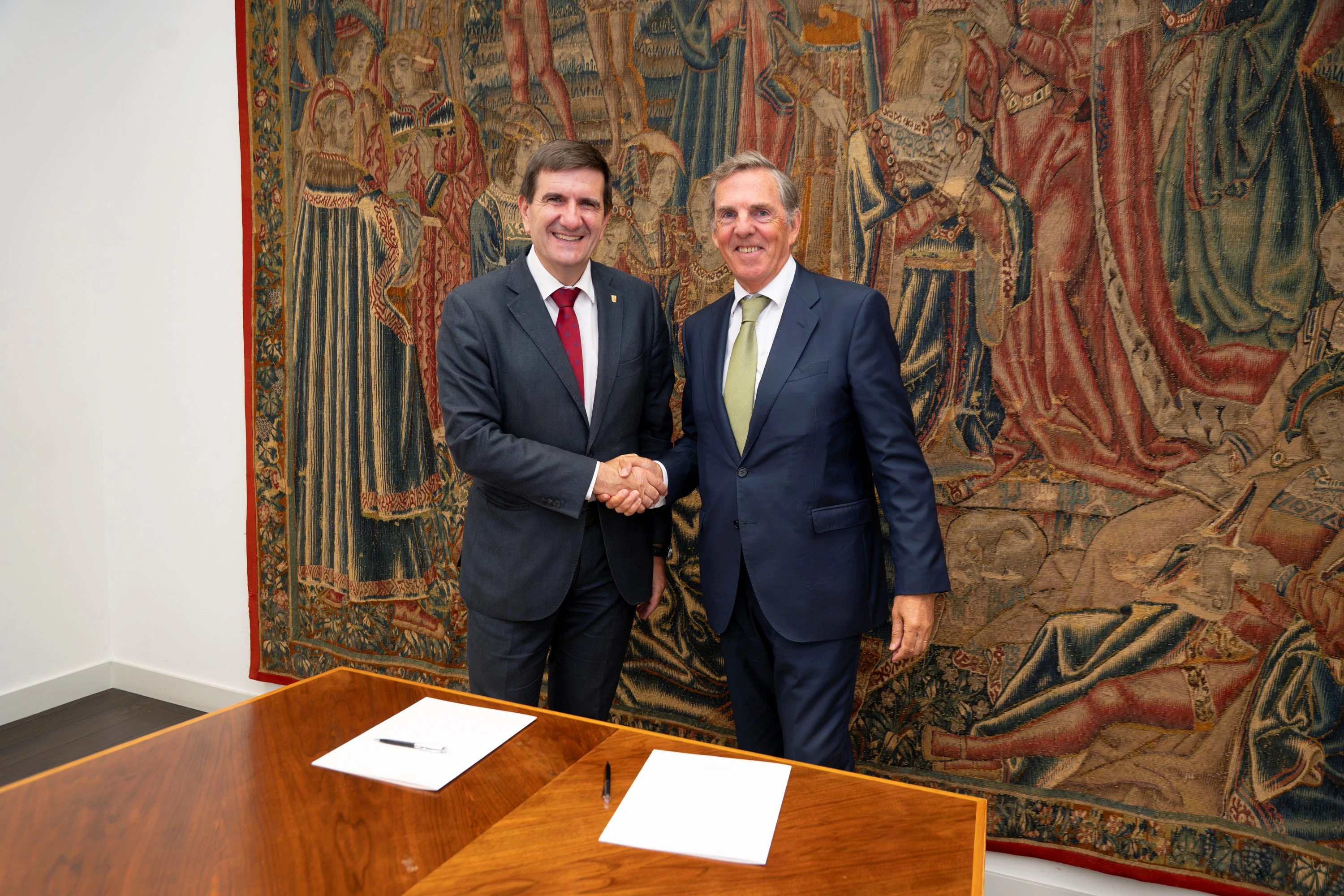 Dos representantes institucionales estrechan la mano junto a una mesa con documentos firmados en un espacio histórico.