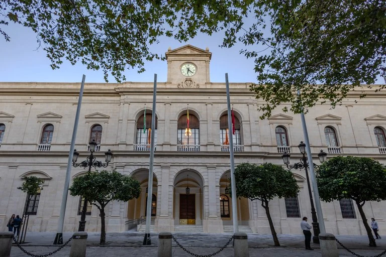 Fachada del Ayuntamiento de Sevilla con arquitectura histórica, árboles y banderas en la entrada principal.