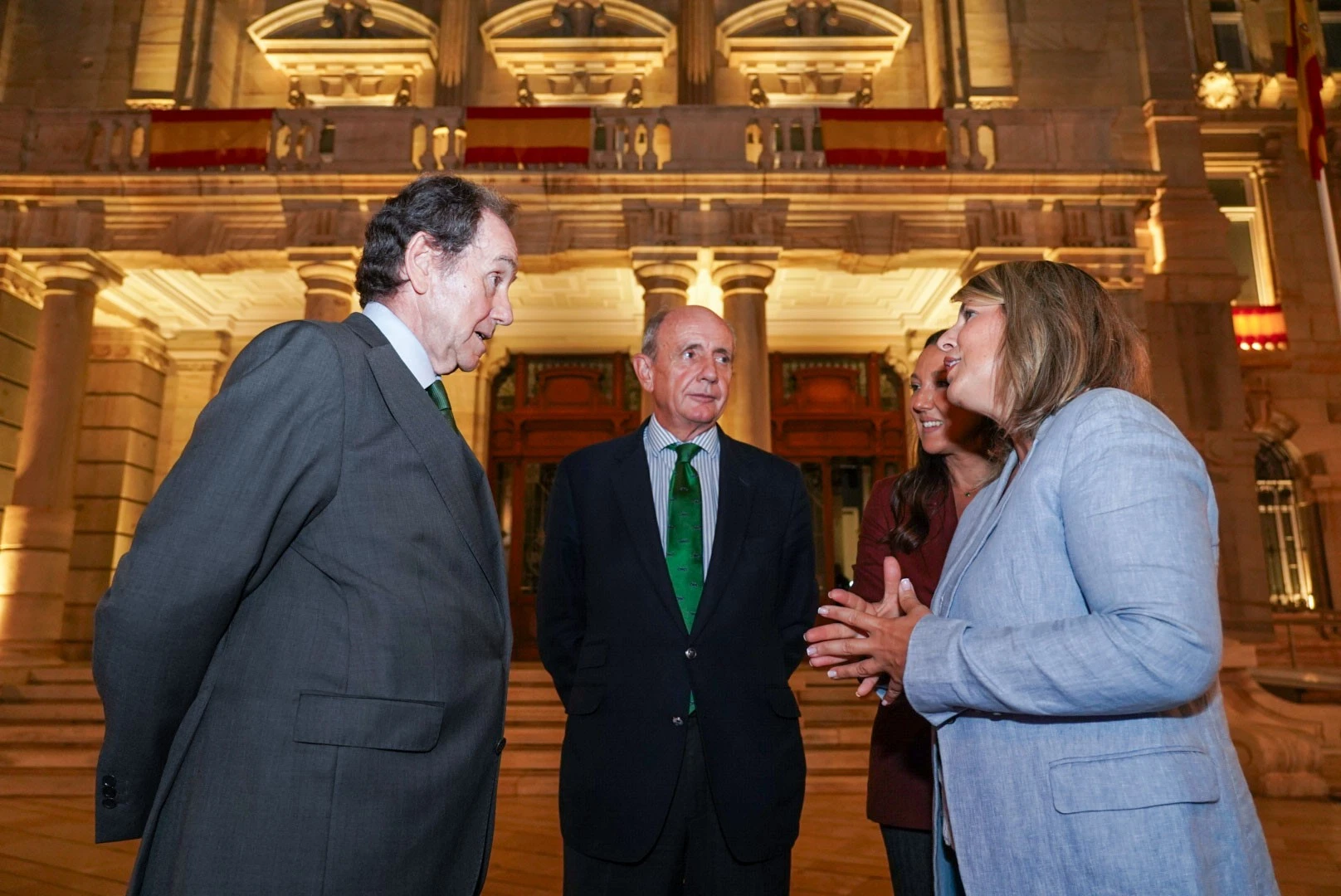 Cuatro representantes institucionales conversan frente a un edificio histórico iluminado y decorado con banderas de España.