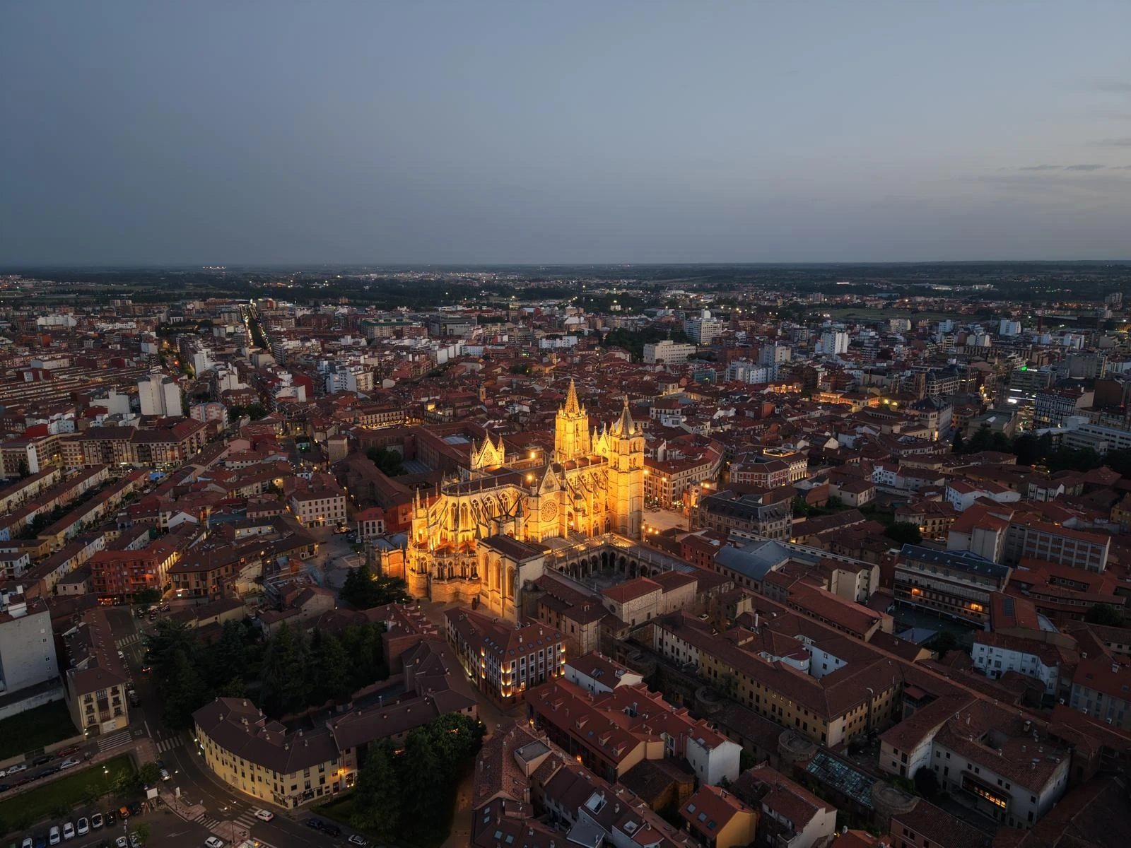 Vista aérea de la catedral de León iluminada al anochecer, rodeada de edificios y calles del centro histórico de la ciudad.