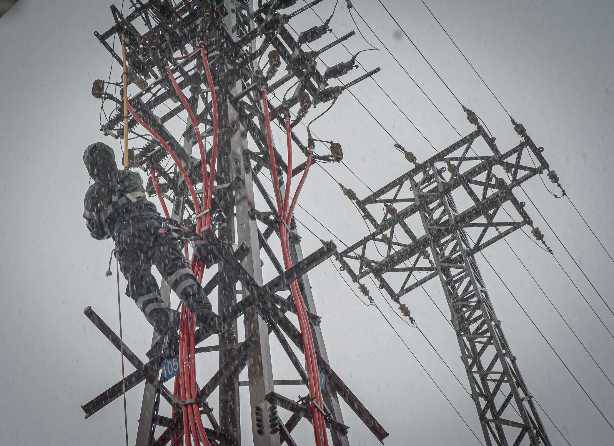 Técnico realizando trabajos en líneas eléctricas de alta tensión en torre de transmisión durante lluvia intensa.