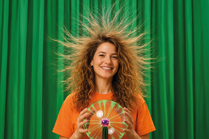 Mujer sonriente con pelo electrizado, camiseta naranja y una esfera transparente con un diseño luminoso de energía sobre fondo verde