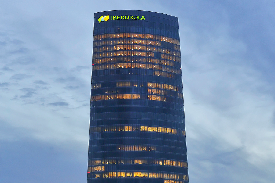 Torre Iberdrola iluminada al atardecer, mostrando sus ventanas encendidas y rodeada de edificios y árboles en primer plano.