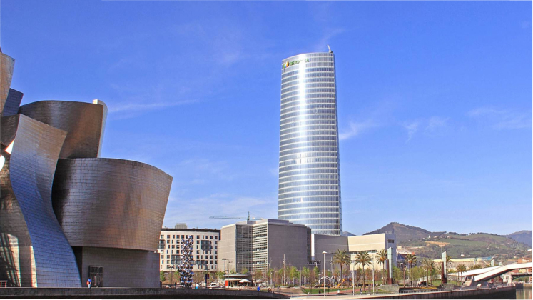 Vista de la Torre Iberdrola en Bilbao, una estructura moderna y alta, con vidrio reflectante que resalta bajo el cielo azul. A la izquierda de la imagen se encuentra el Museo Guggenheim de Bilbao. En primer plano, se observan palmeras y un paisaje urbano que rodea los edificios.