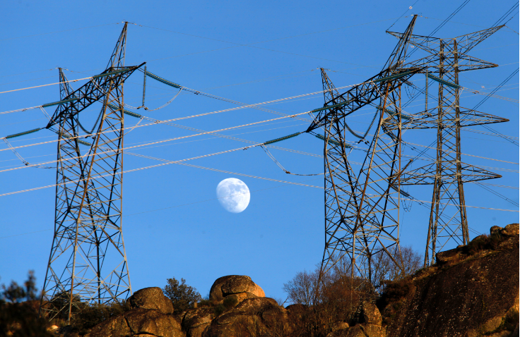Imagen de dos torres de alta tensión eléctricas en primer plano, con cables suspendidos entre ellas. En el fondo, se ve el cielo azul claro y la luna llena parcialmente visible entre las estructuras de las torres.
