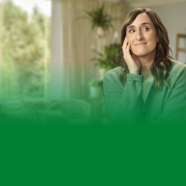 Una mujer sonríe mientras está sentada, mirando hacia un lado con una expresión pensativa y relajada. Lleva un suéter verde y está en un ambiente hogareño con plantas y luz natural en el fondo. El lado izquierdo de la imagen tiene un color verde difuso, lo que podría indicar un diseño de banner o una transición hacia otro contenido visual.