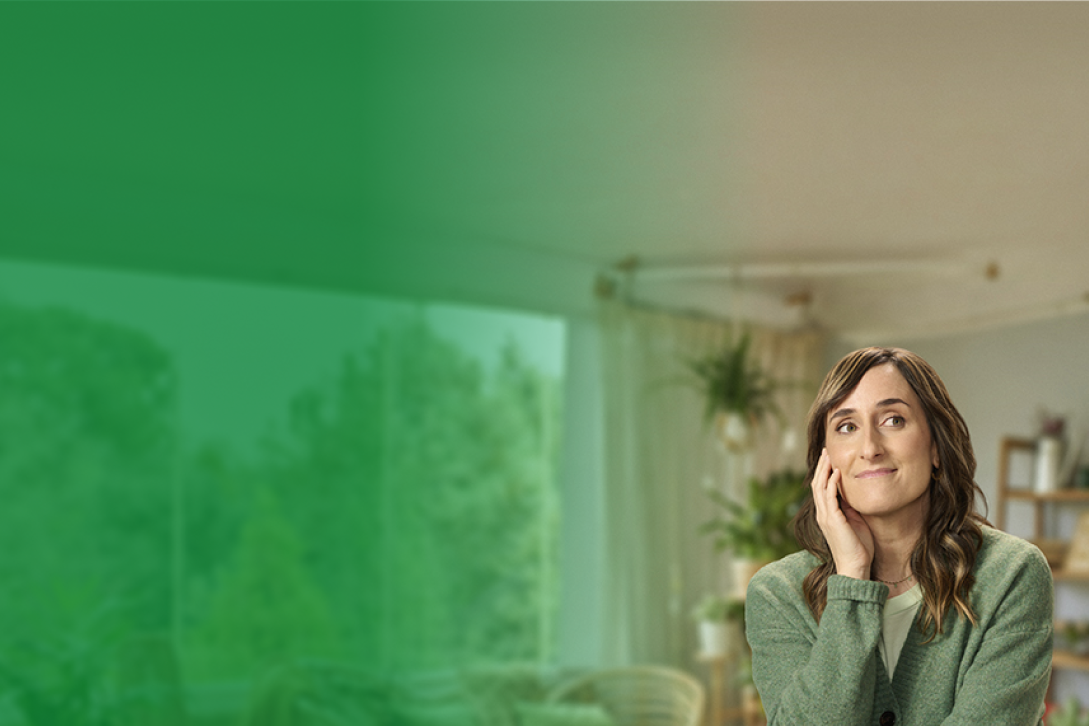 Una mujer sonríe mientras está sentada, mirando hacia un lado con una expresión pensativa y relajada. Lleva un suéter verde y está en un ambiente hogareño con plantas y luz natural en el fondo. El lado izquierdo de la imagen tiene un color verde difuso, lo que podría indicar un diseño de banner o una transición hacia otro contenido visual.