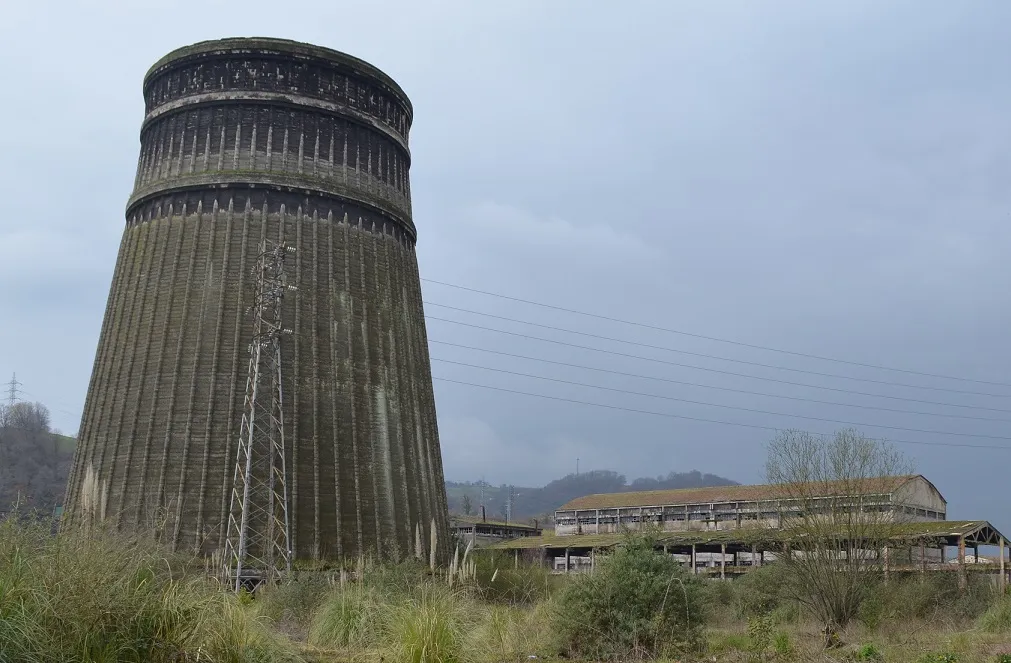 Former cooling tower alongside industrial facilities and power lines in a natural setting.
