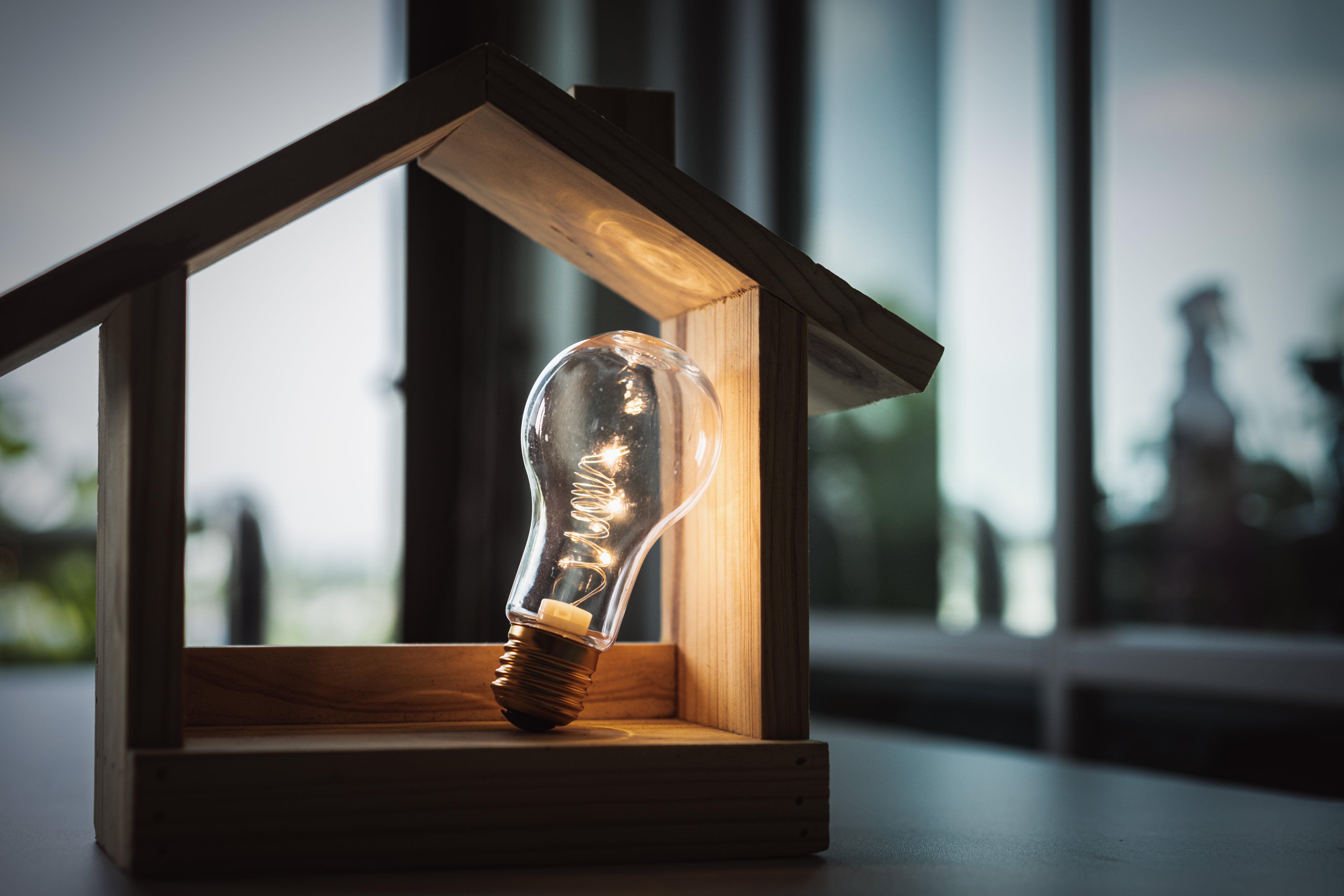 A lit lightbulb inside a small wooden house. The light from the bulb creates a contrast with the dark background and the slightly blurred environment of a window with natural light.